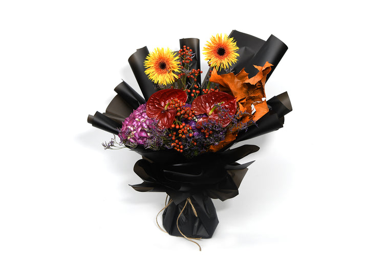 A bouquet of flowers including golden gerberas, an chocolate anthurium, a purple hydrangea, and other mixed flowers, wrapped in black paper.